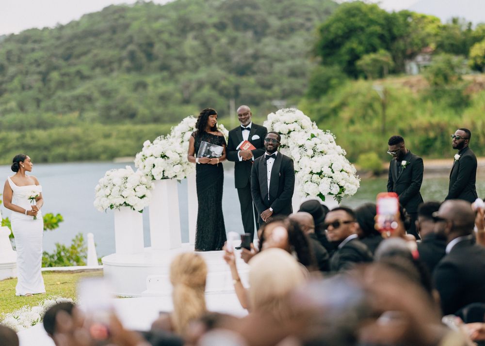 Wedding ceremony takes place outdoors with white chairs arranged on green lawn overlooking scenic lake and hills.