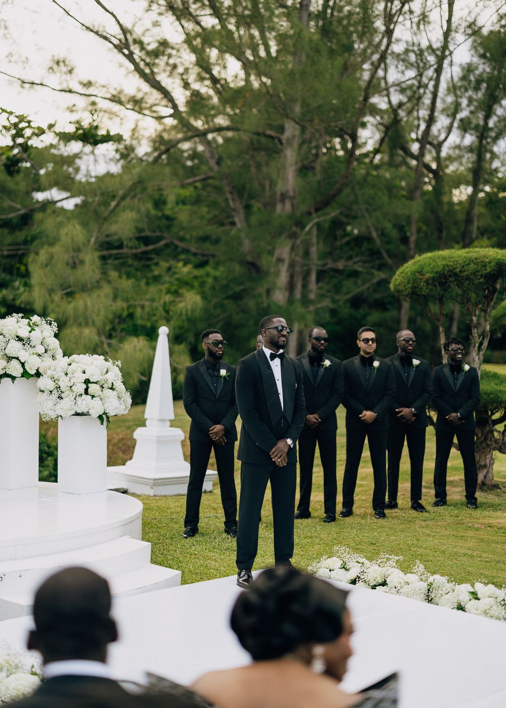 An elegant outdoor wedding ceremony setup with black-suited groomsmen standing on a green lawn near white floral decorations.