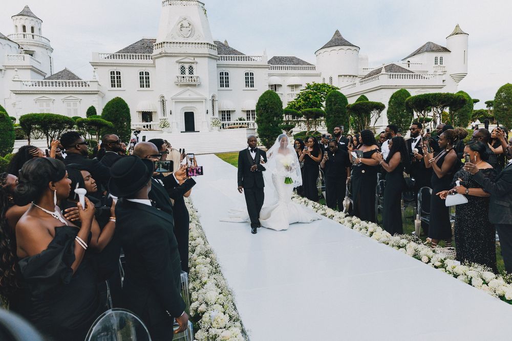 A panoramic view of a wedding ceremony with guests in formal black attire gathered around white floral decorations.