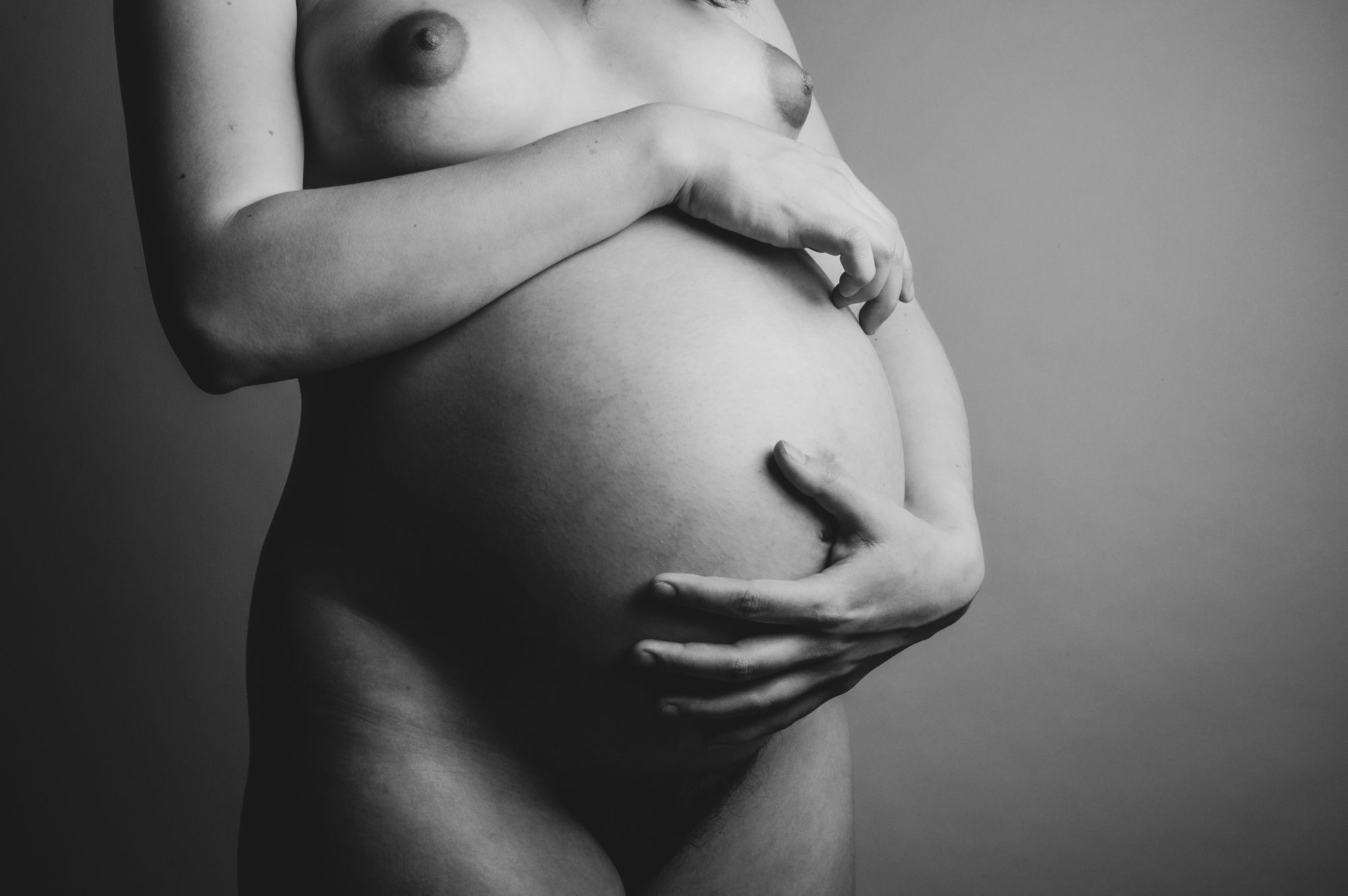 Zwartwit detailportret van een zwangere buik met handen die zacht ondersteunen, tijdloos en elegant gefotografeerd in de studio.