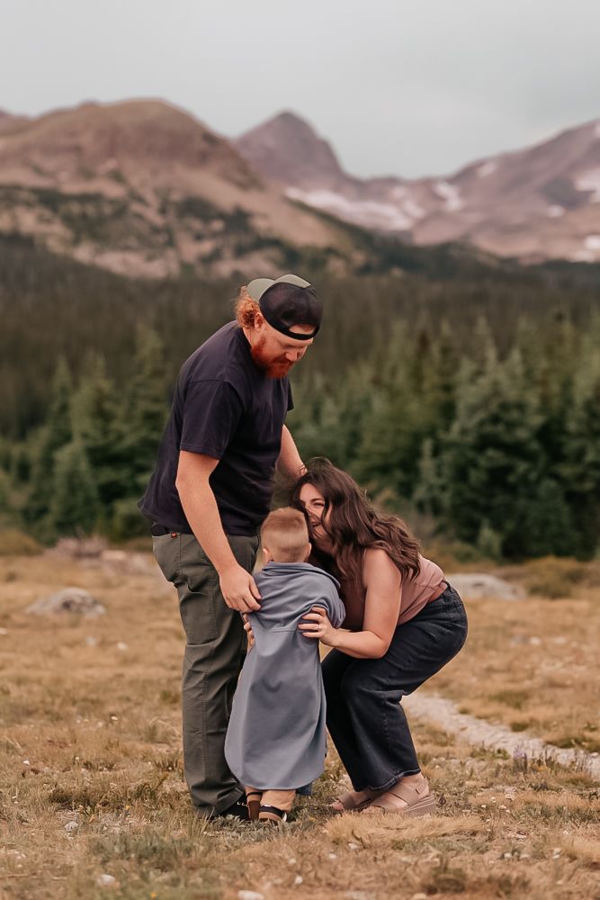 A couple shares a tender moment in a scenic mountain meadow with evergreen forest and peaks in the background.
