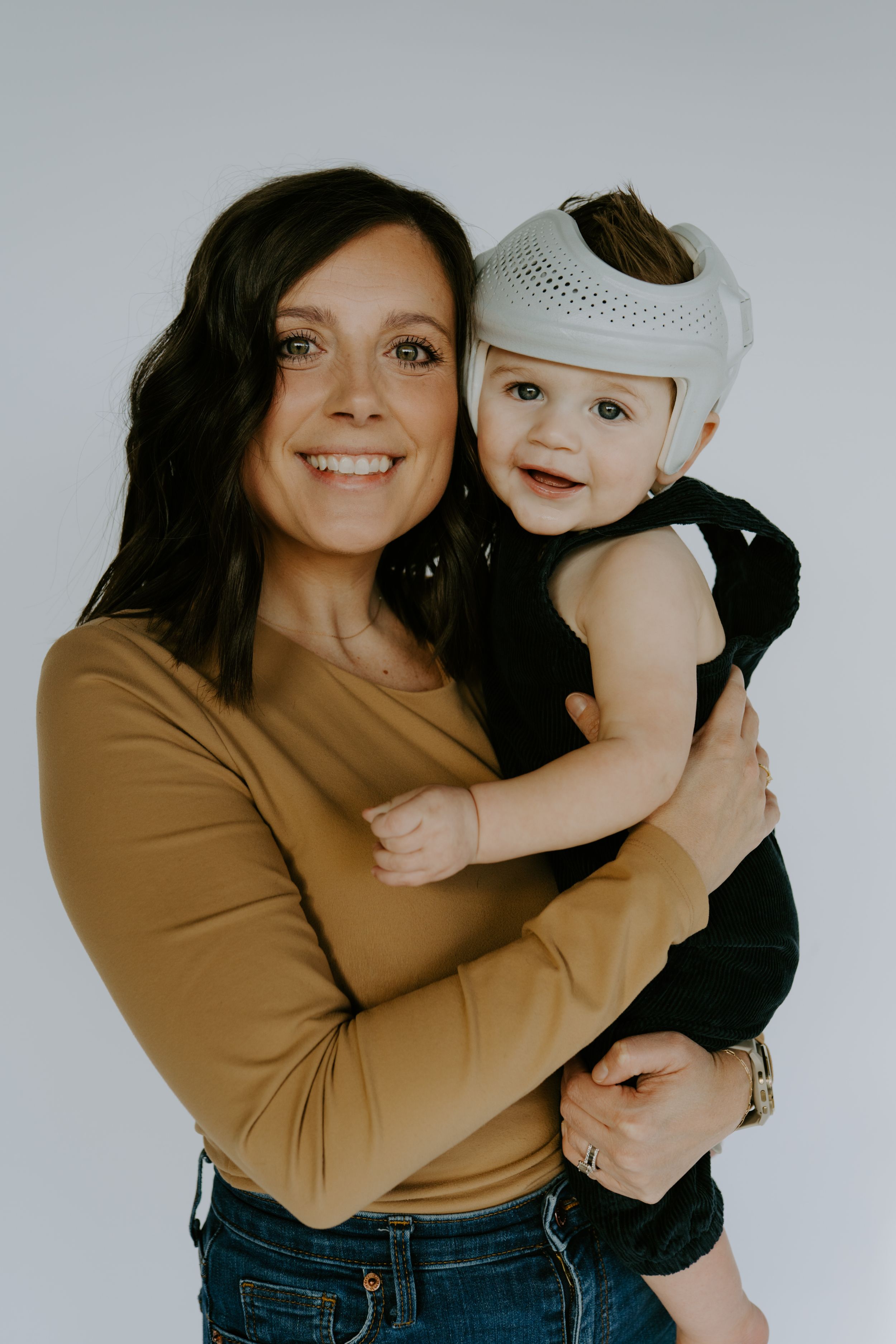 Mother in tan sweater holds baby in black outfit against white backdrop.
