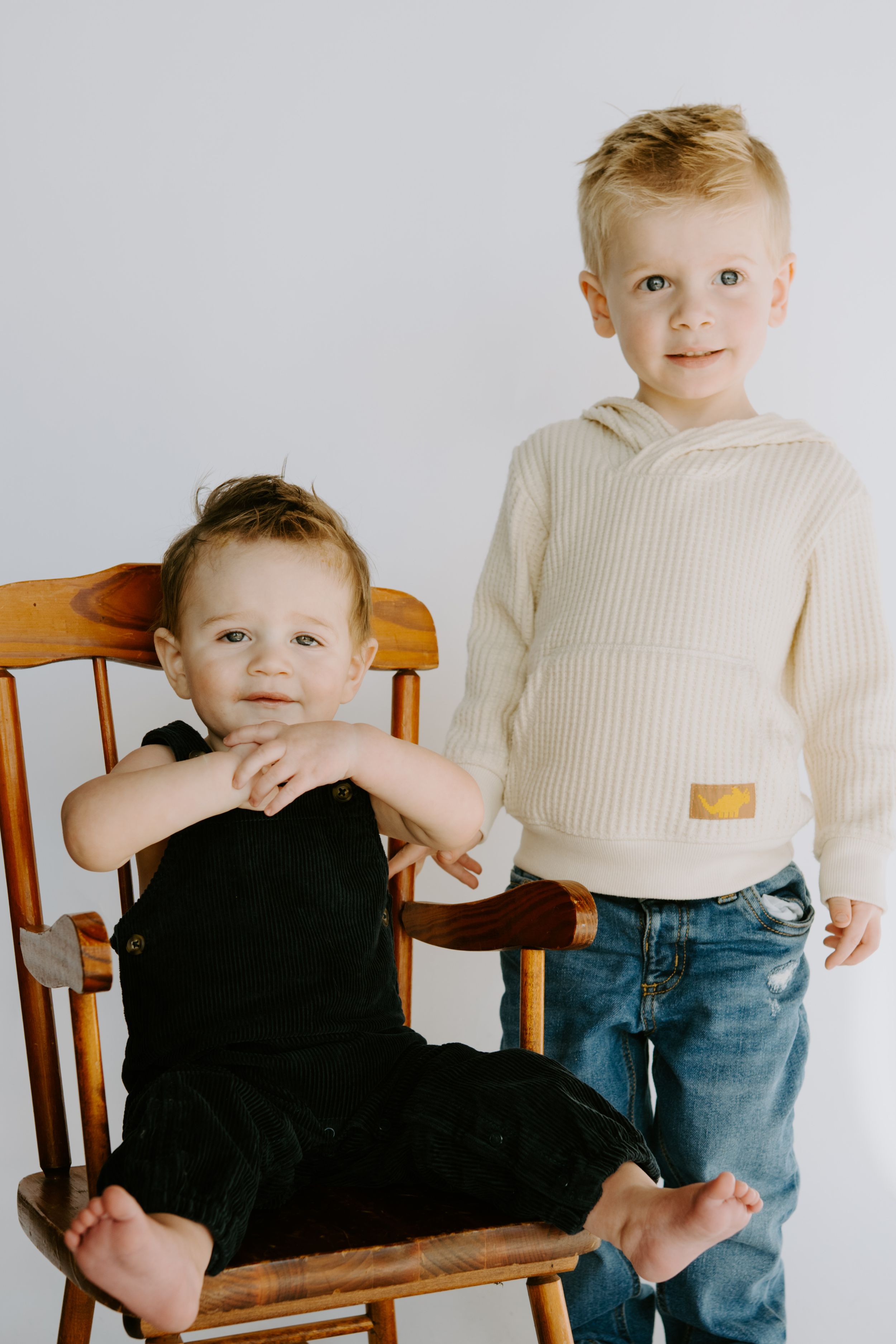 Two children in cream sweaters pose on wooden rocking chair.