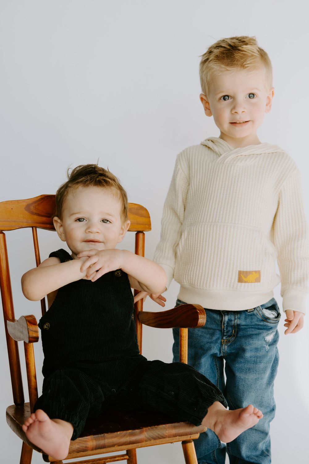 Two children in cream sweaters pose on wooden rocking chair.