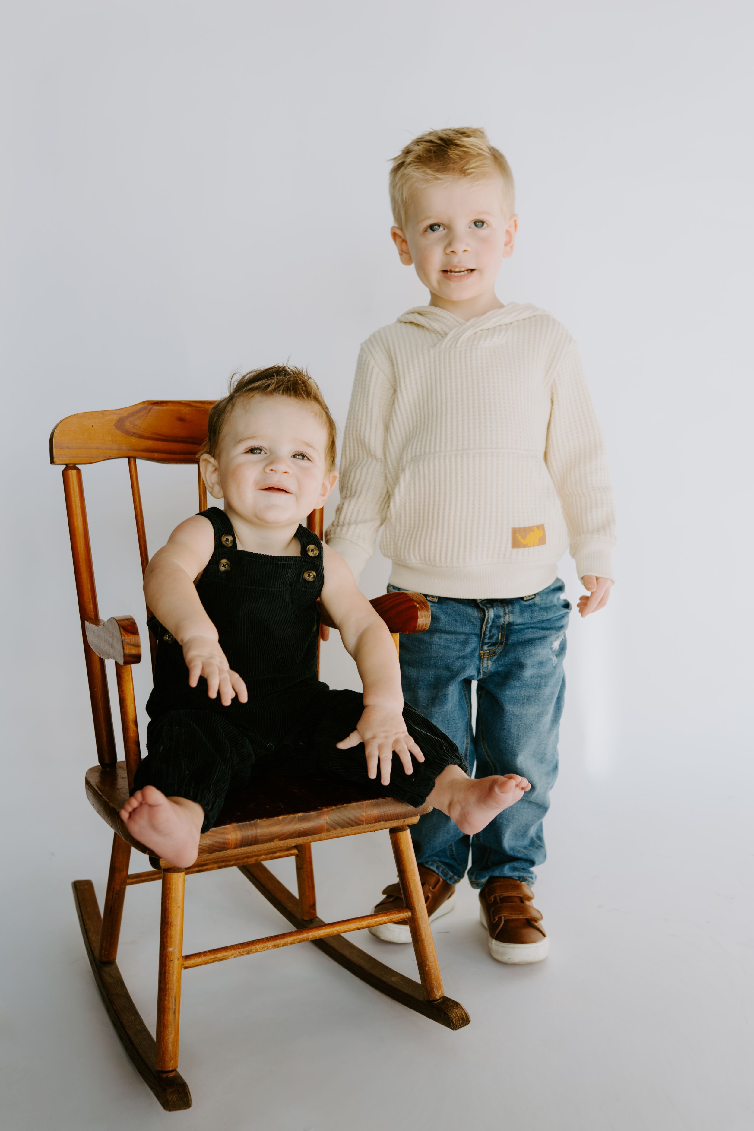 Two children pose with wooden rocking chair in minimalist studio setting.