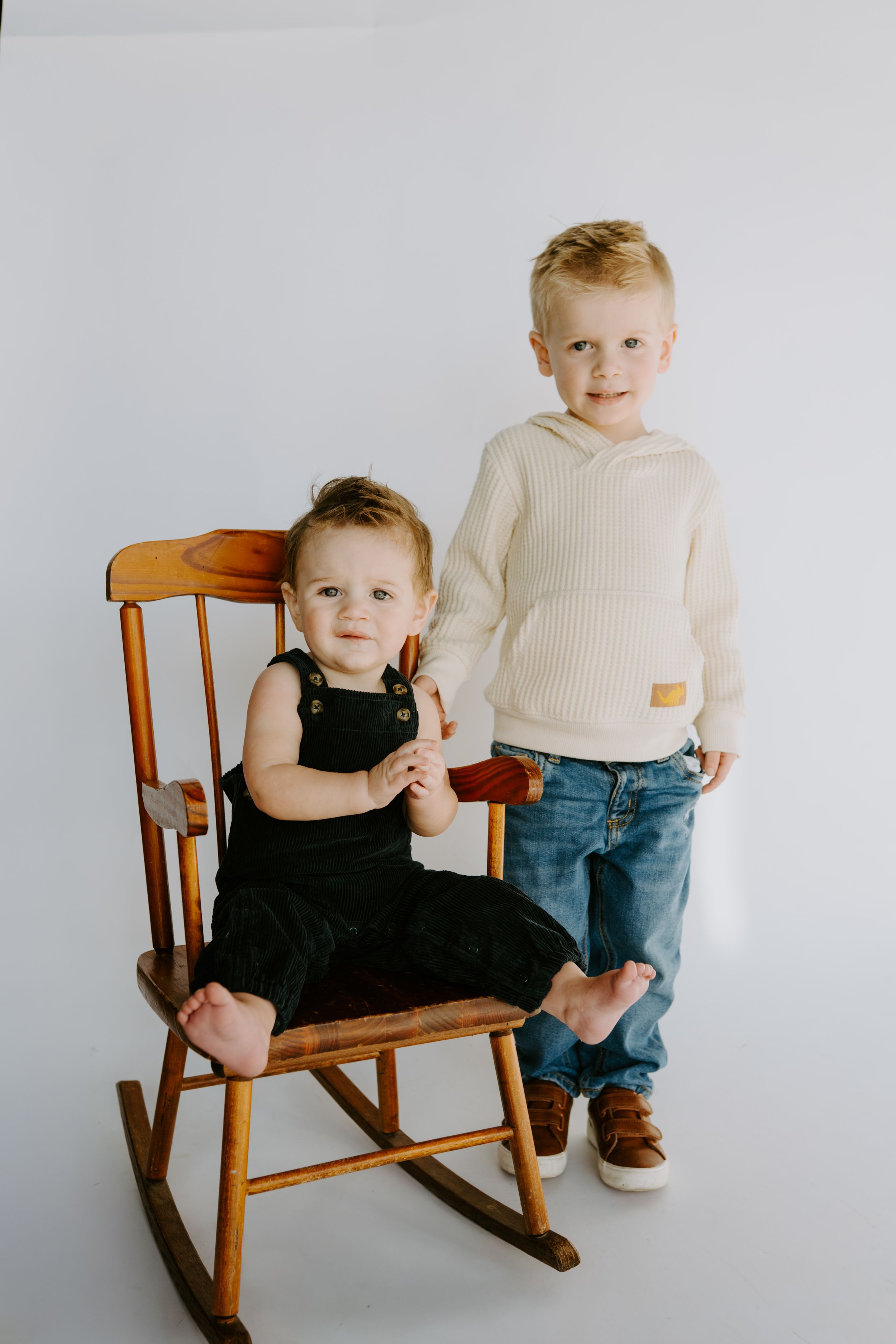 Children in casual wear pose with vintage wooden rocking chair against white backdrop.