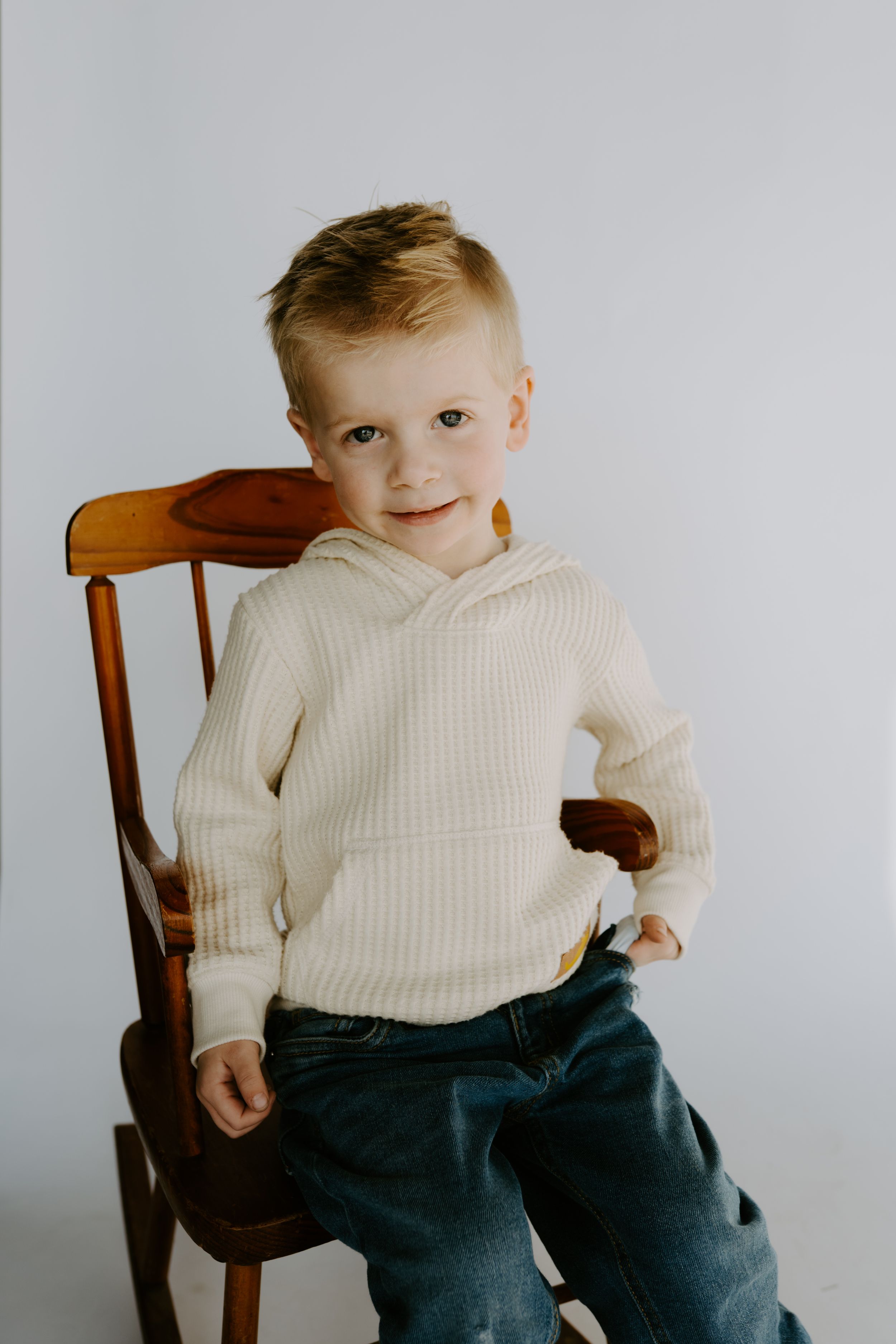 Multiple angles of a toddler wearing a white knit sweater and dark pants seated in a wooden chair.