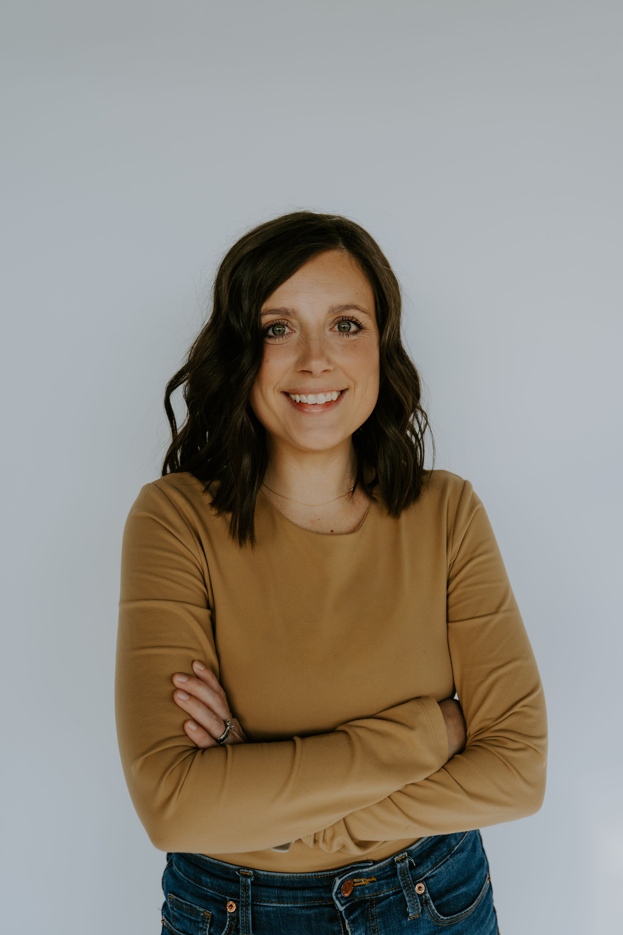 A woman in a brown sweater and jeans poses casually against a white background.