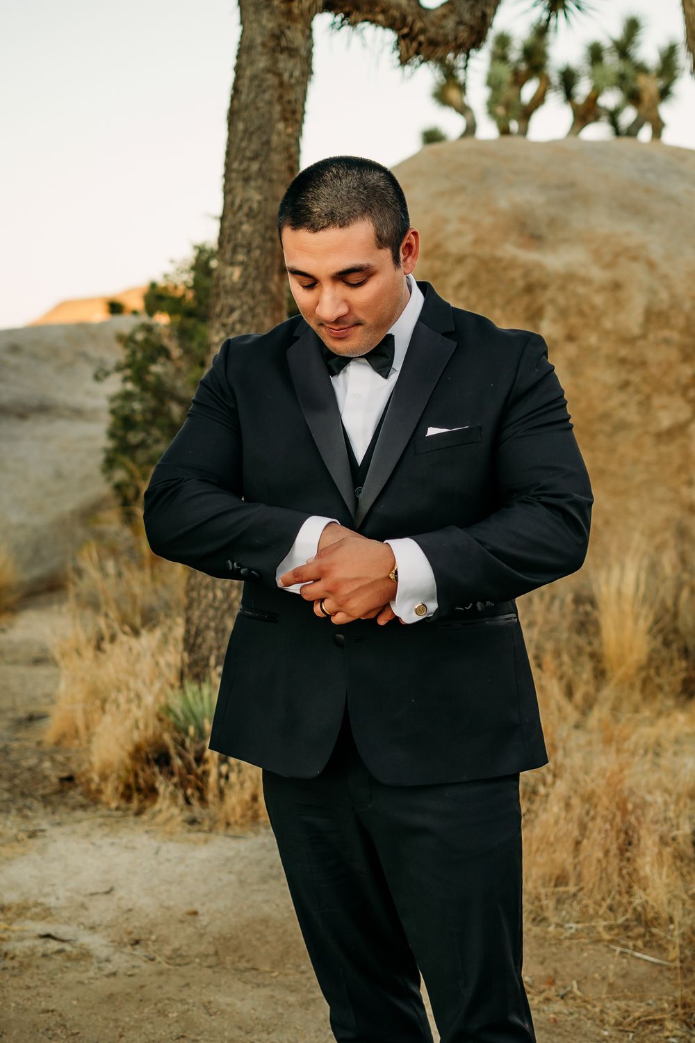 A groom adjusts his formal attire while standing in an outdoor desert setting.