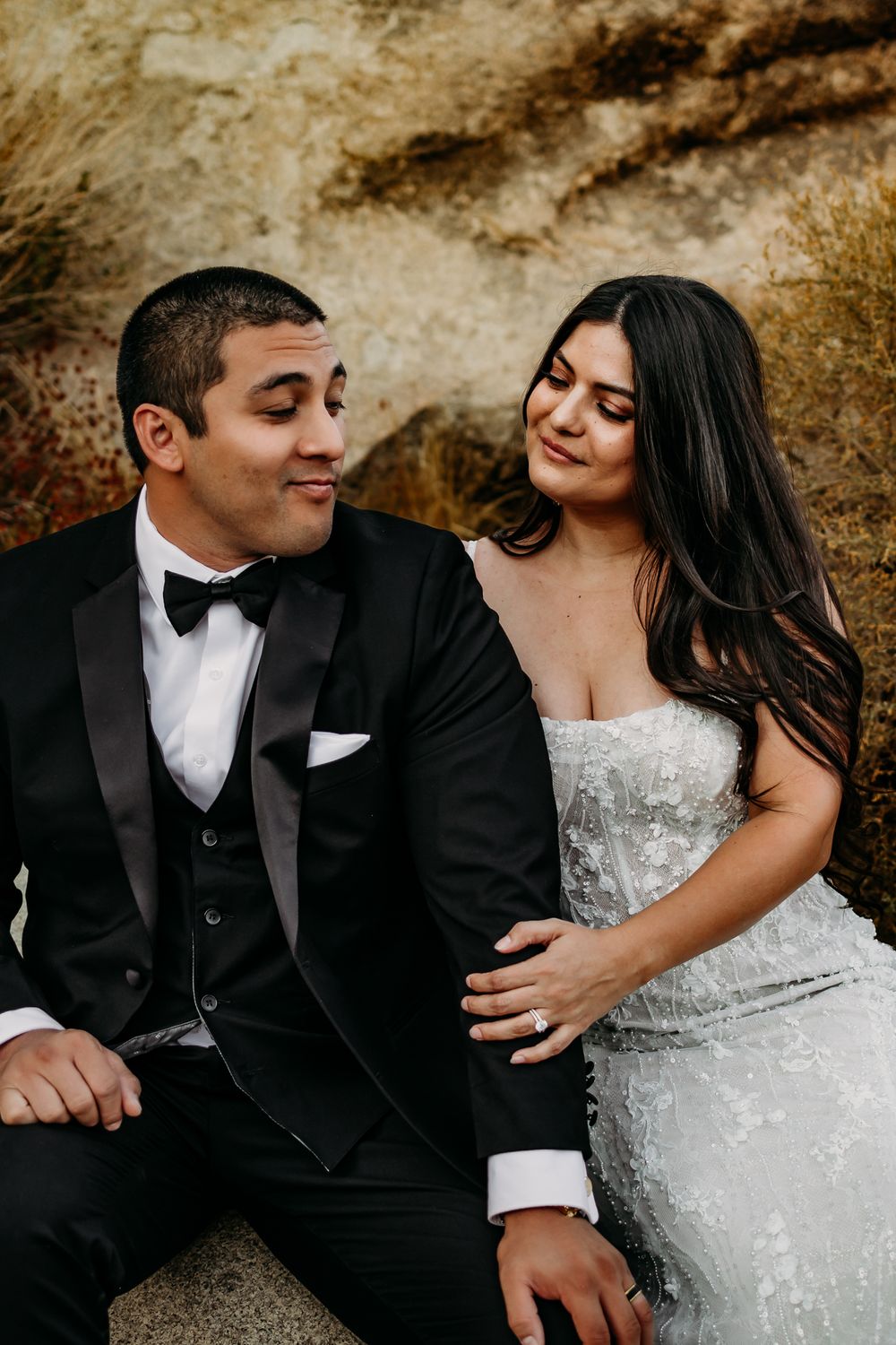 A romantic wedding couple in formal attire share intimate moments against a textured rock wall backdrop.