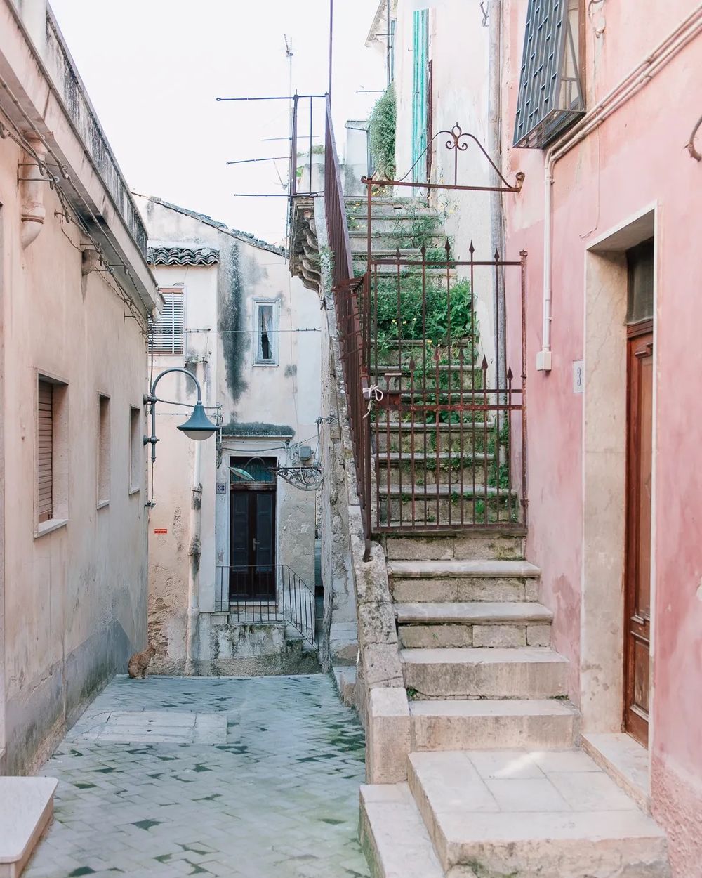 A narrow Mediterranean street with pastel walls and an outdoor staircase leading to an upper floor.
