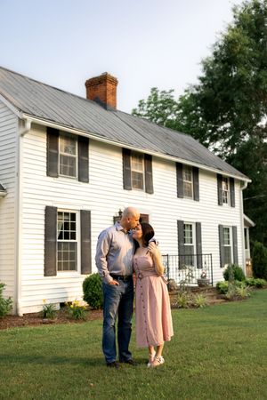 Couple poses romantically in front of a classic white farmhouse with brown shutters.