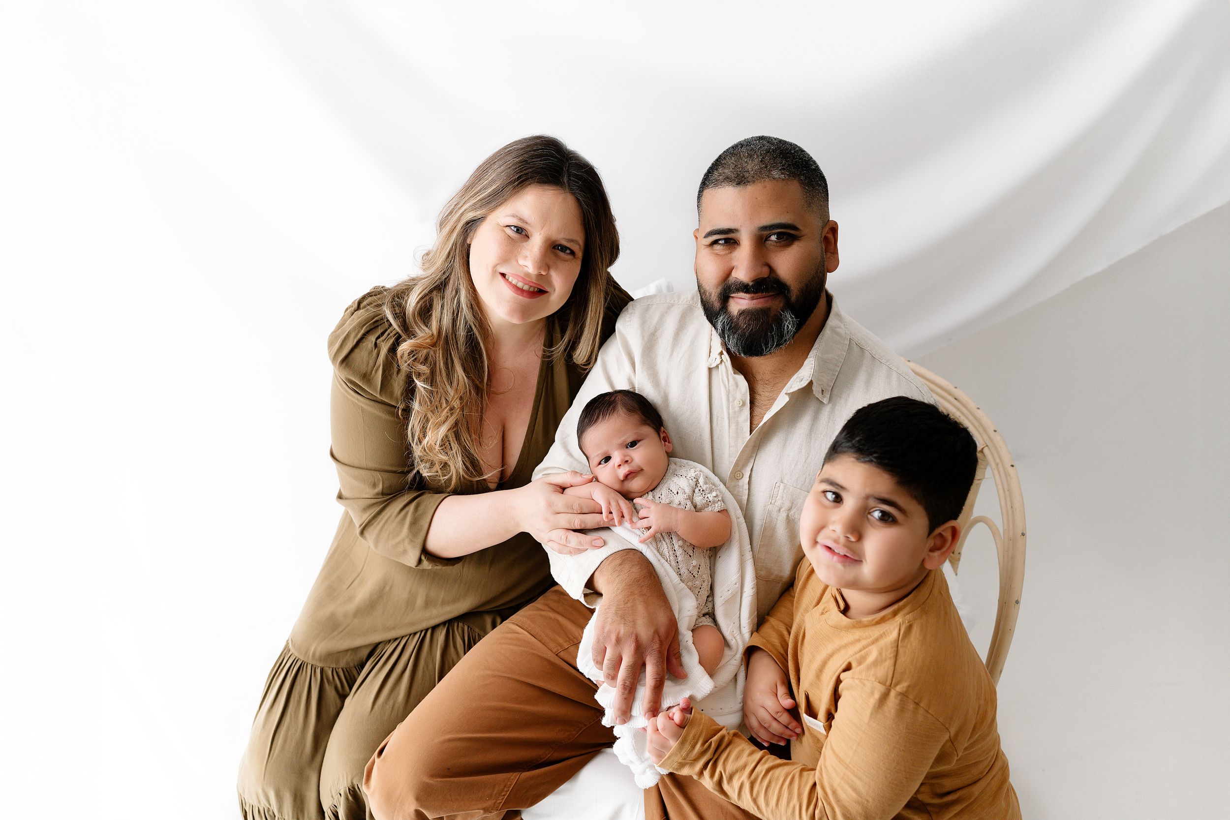 A family poses together in a warm neutral-toned studio setting wearing coordinated brown and beige outfits.