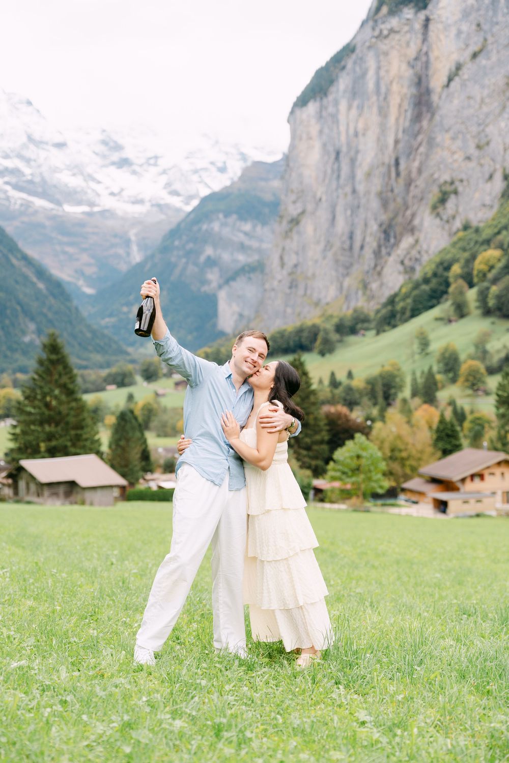 Playful couple dancing and twirling in a green meadow with dramatic Swiss Alps mountains and chalets in background.