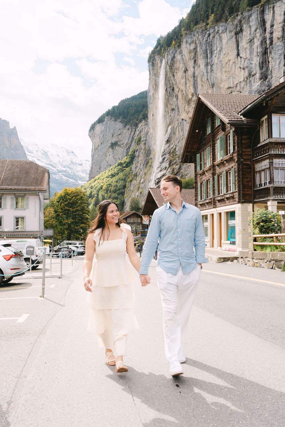 A couple strolls along a scenic mountain road in Lauterbrunnen, Switzerland with dramatic cliffs and traditional chalets in the background.