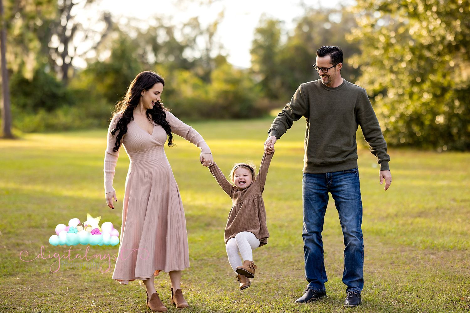A family holds hands and plays together in a sunny park setting with autumn trees in the background.