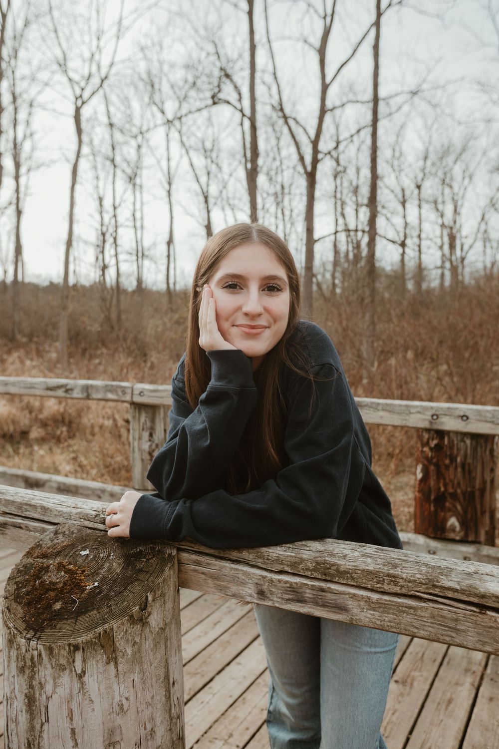 A person in a black sweater leans against wooden railings on a deck surrounded by bare winter trees in the background.
