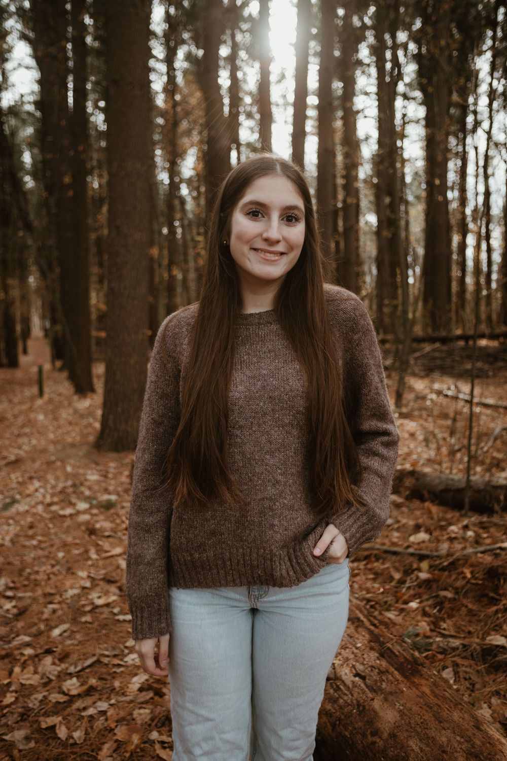 Person wearing brown sweater and light jeans stands in autumn forest with fallen leaves on ground.