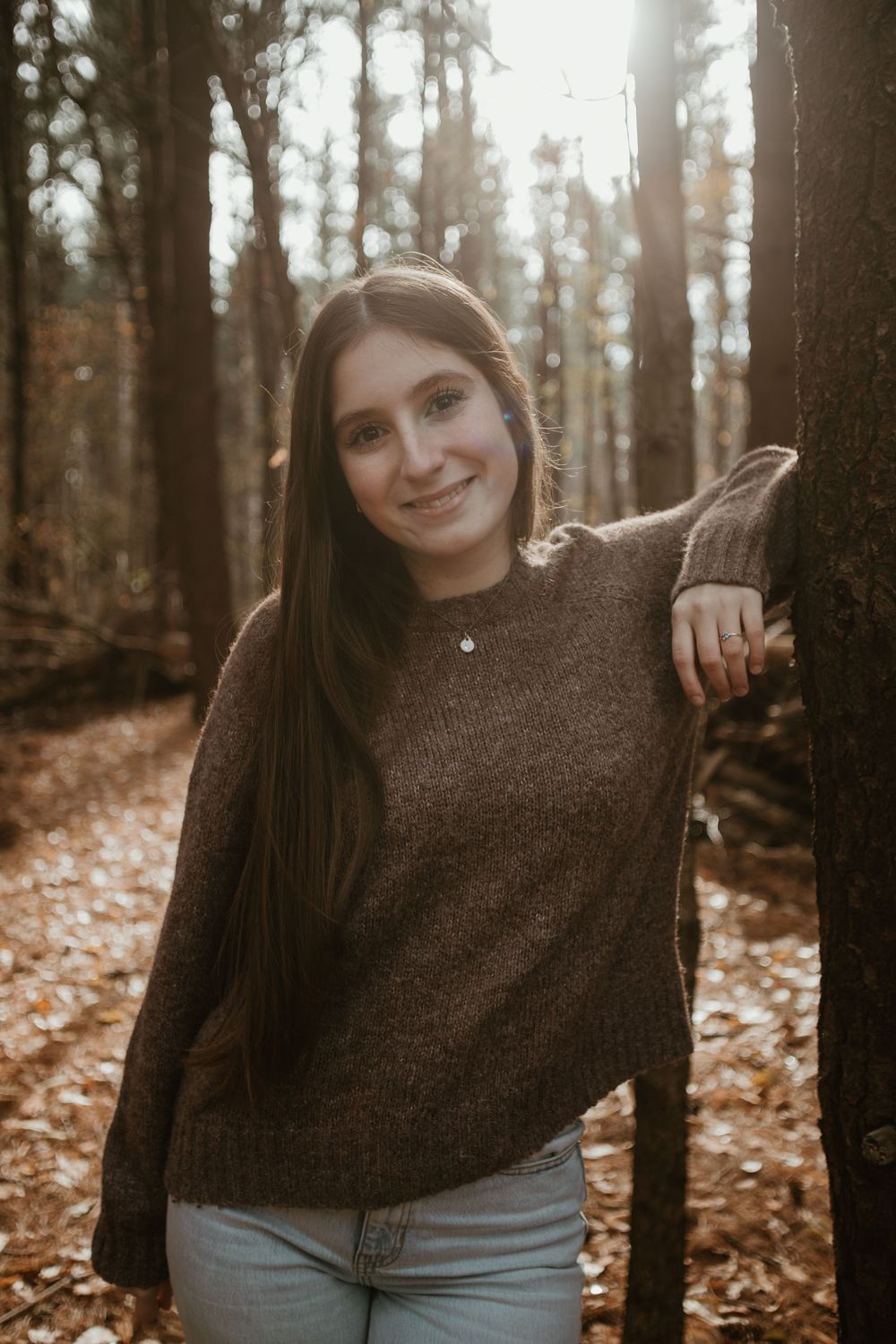 A person in a brown sweater poses playfully in an autumn forest with fallen leaves on the ground.