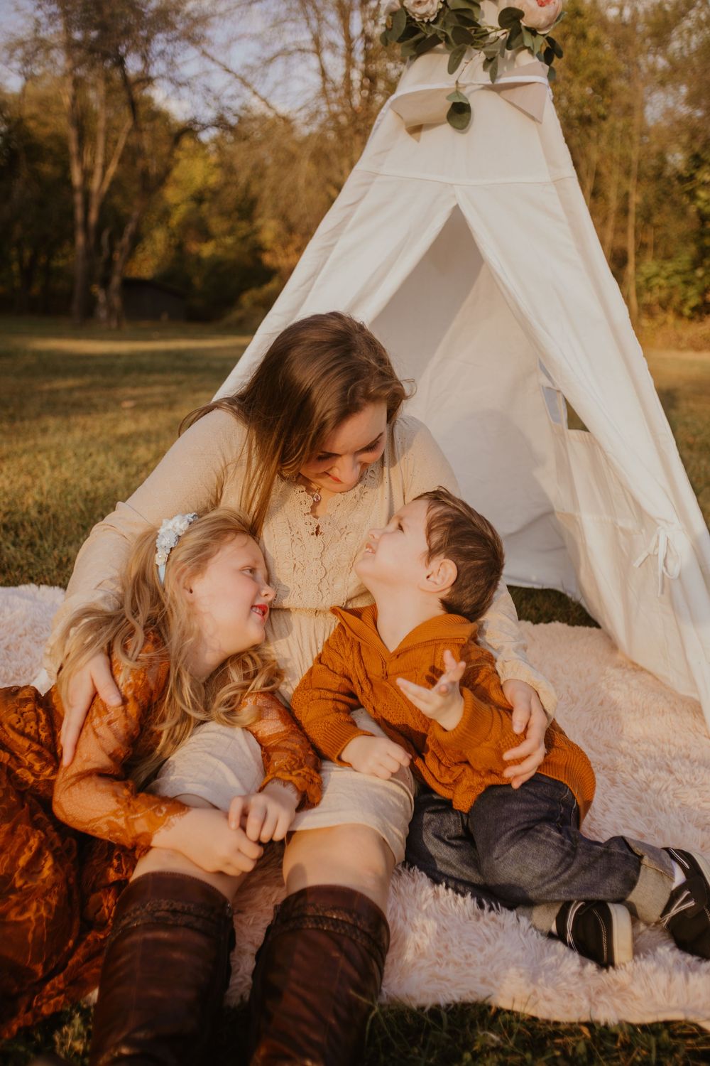 A cozy scene unfolds near a white teepee tent with people sharing tender moments on a blanket in warm autumn light.