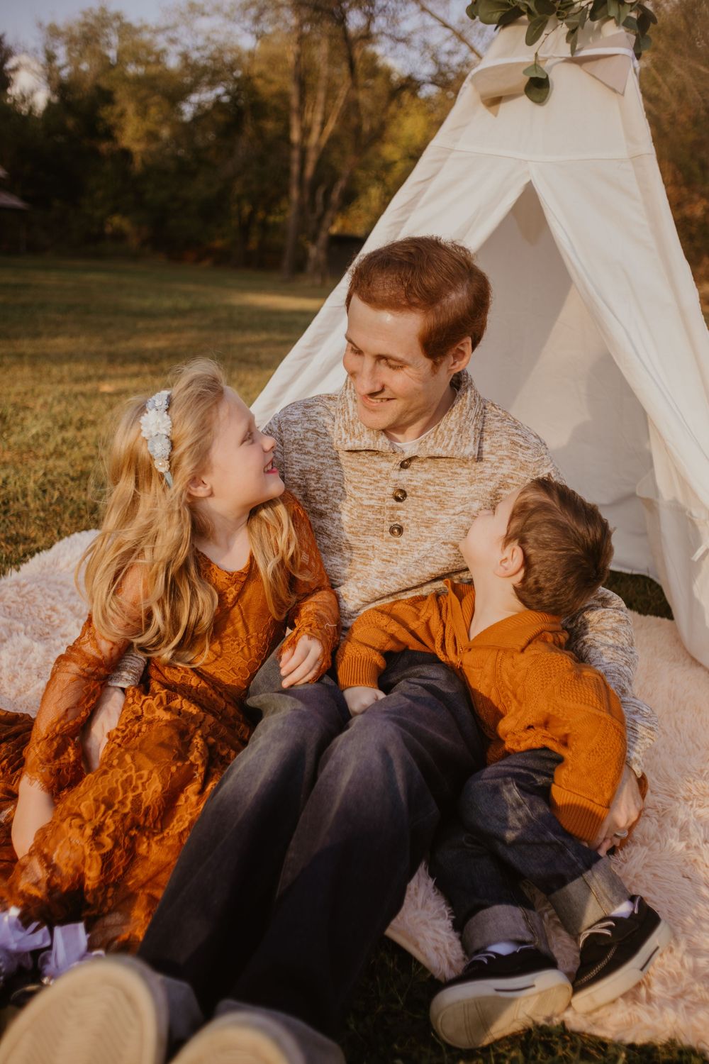 Family sharing tender moments together while sitting near a white teepee tent at sunset in an outdoor setting.