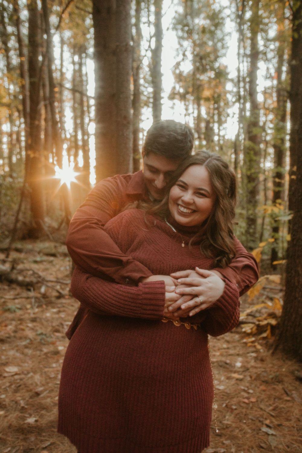 A couple embraces warmly in a sunlit autumn forest wearing matching burgundy sweaters.