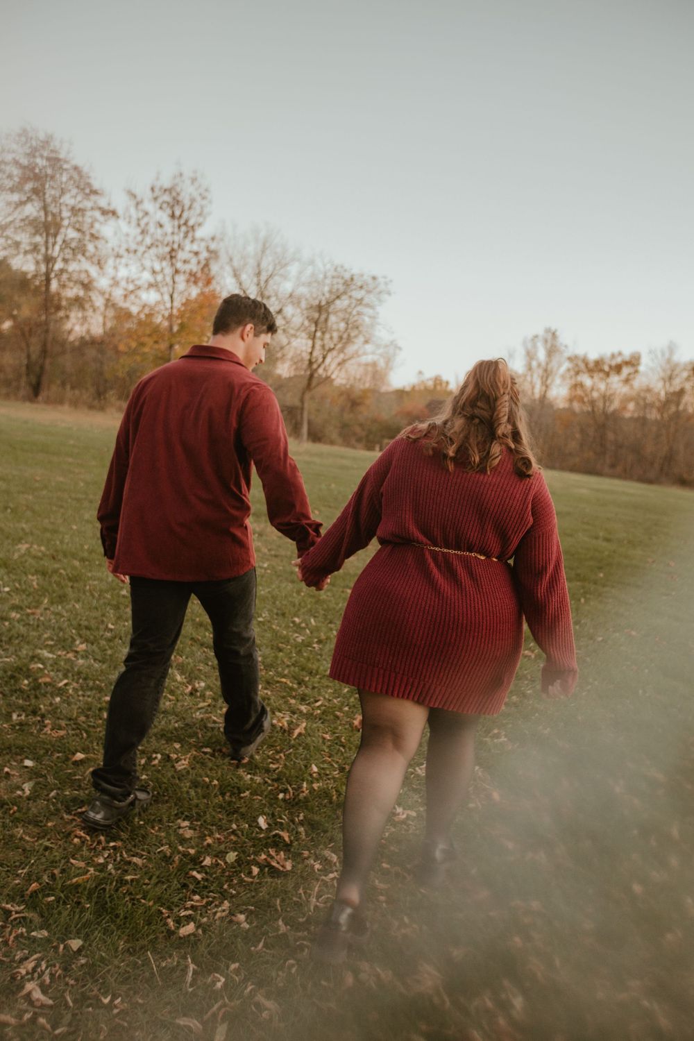 Two people in burgundy sweaters walk together through a misty autumn field with fallen leaves at sunset.