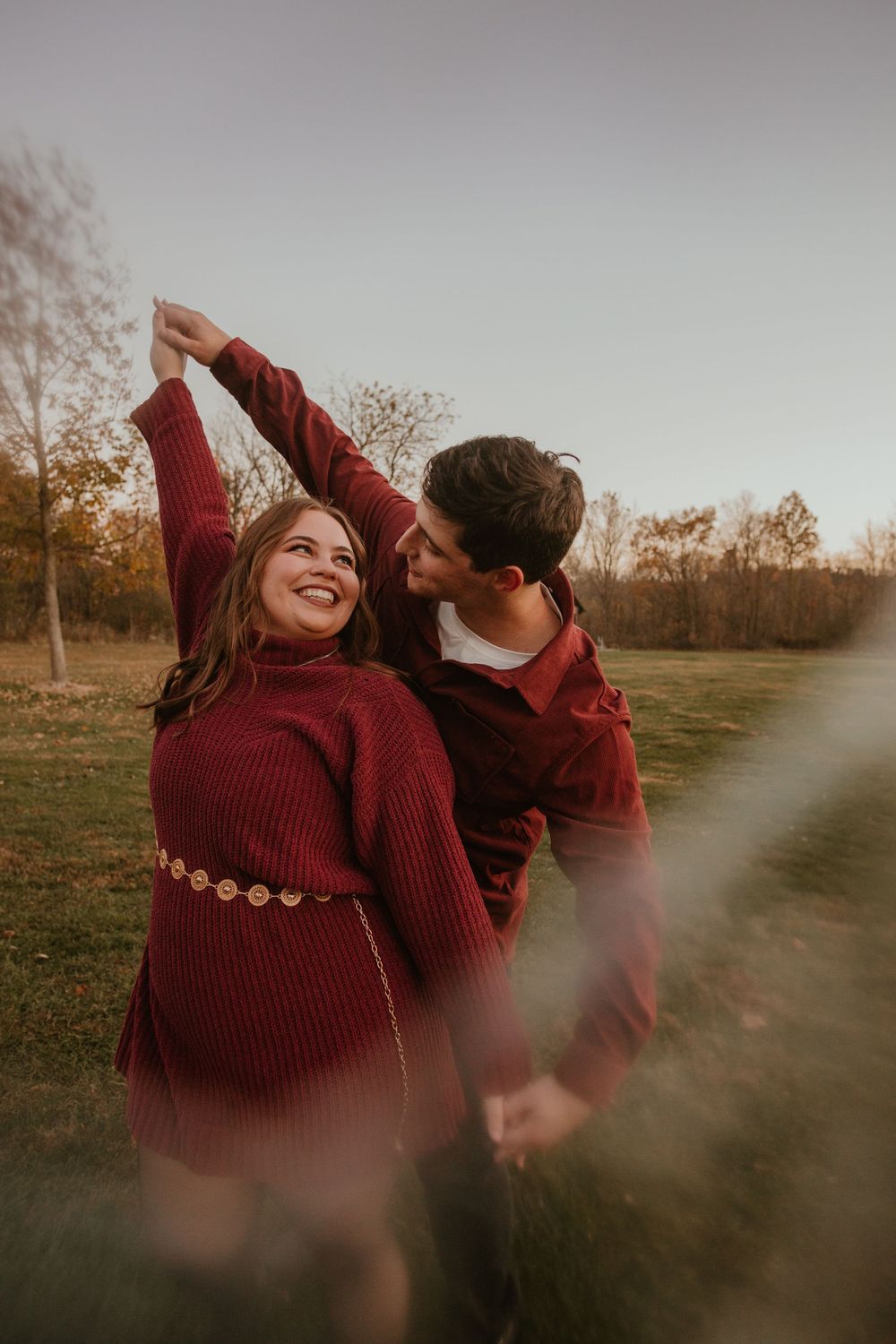 A couple in matching burgundy sweaters share intimate moments in a golden autumn field at sunset.