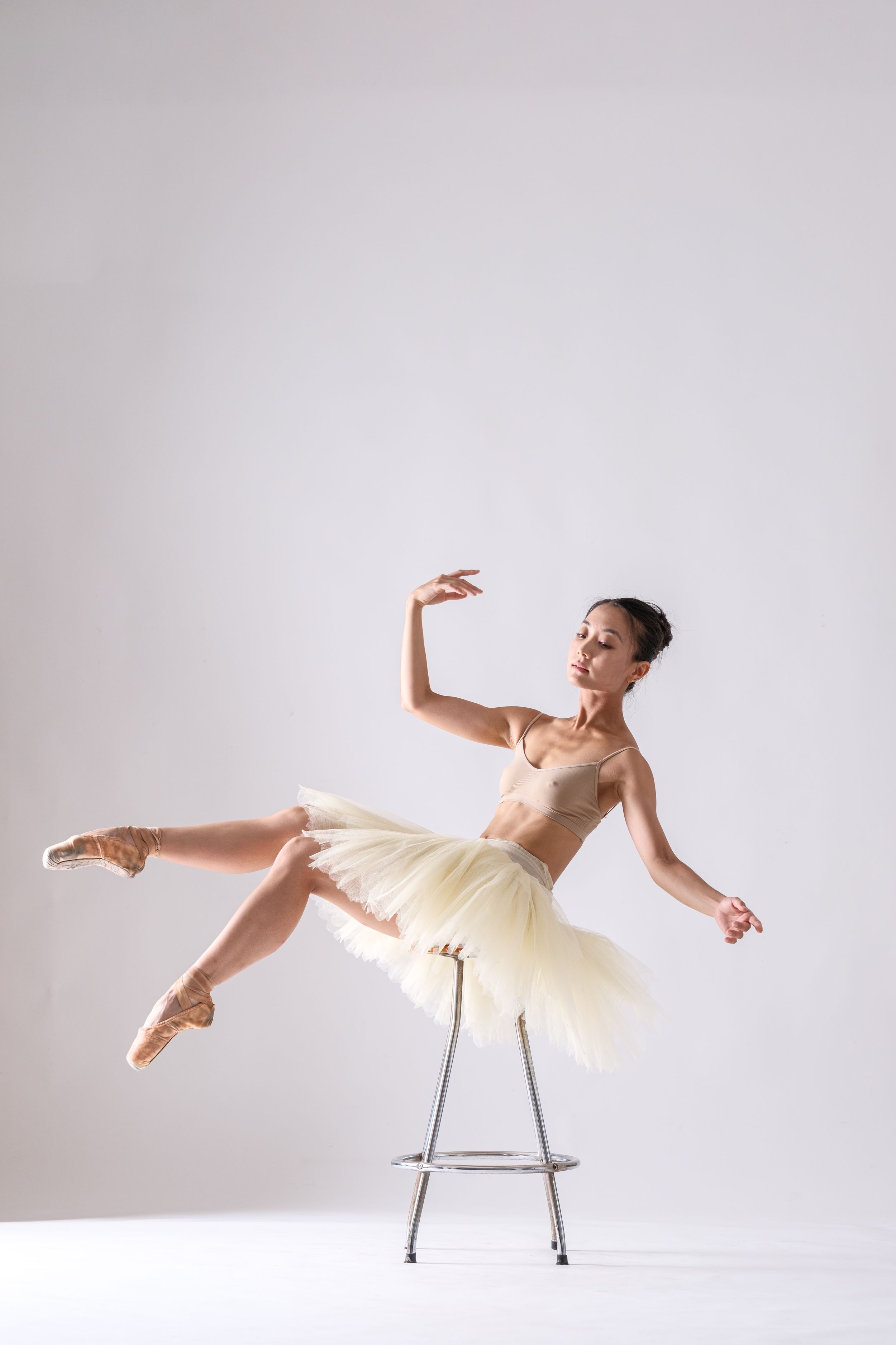 Dancer in cream tutu creates elegant poses while incorporating metal stool in bright studio.