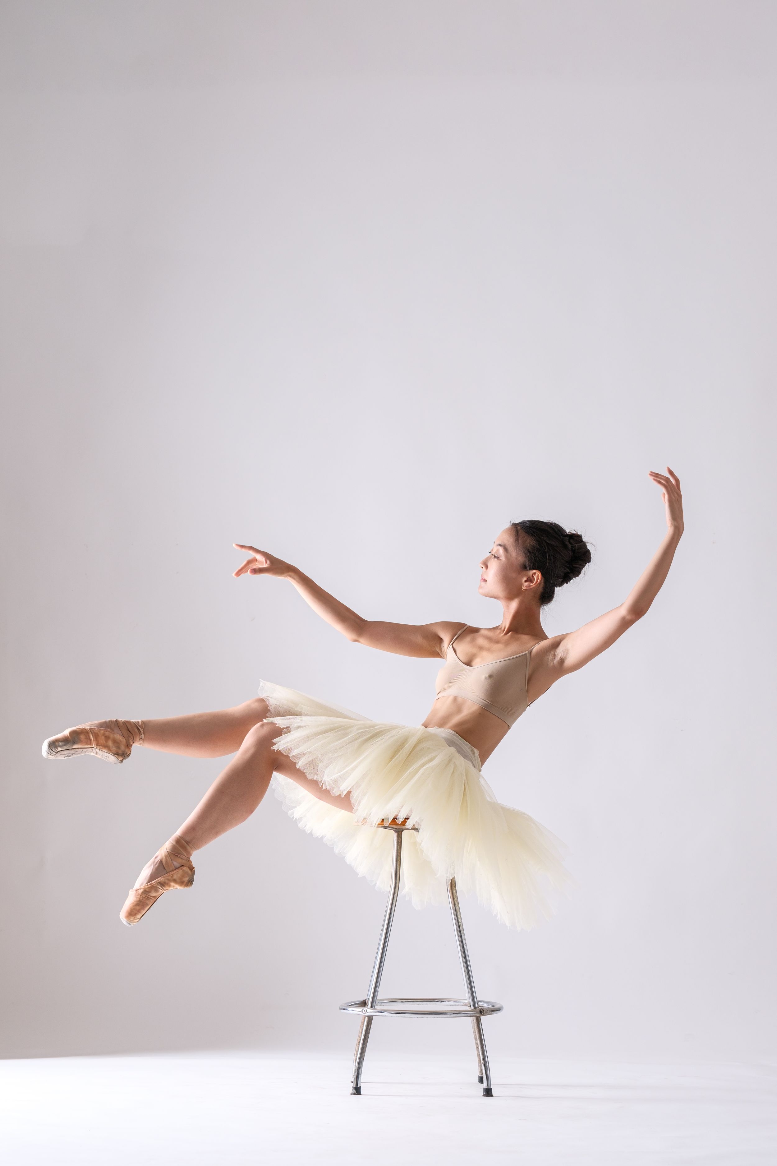 Ballerina in cream tutu performs graceful pose while balanced on metal chair against white backdrop.