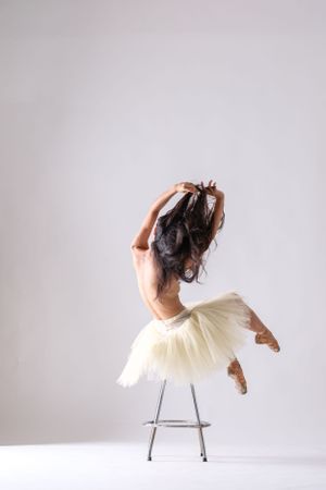 Contemporary dancer in cream tutu executes dynamic pose using metal chair in minimalist setting.
