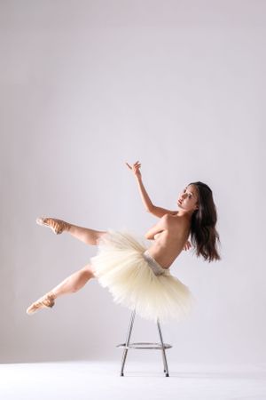 Dancer creates dramatic lines while balanced on chair in cream tutu against stark white backdrop.