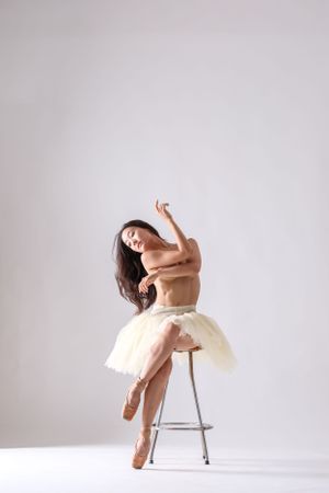Artistic dance pose featuring performer in cream tulle dress using metal chair as support.