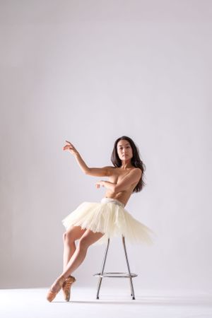 Dynamic dance pose featuring cream tutu and metal chair in clean white studio environment.
