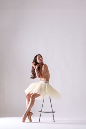 Ballet dancer in cream tutu performs graceful poses with a metal stool against white backdrop.