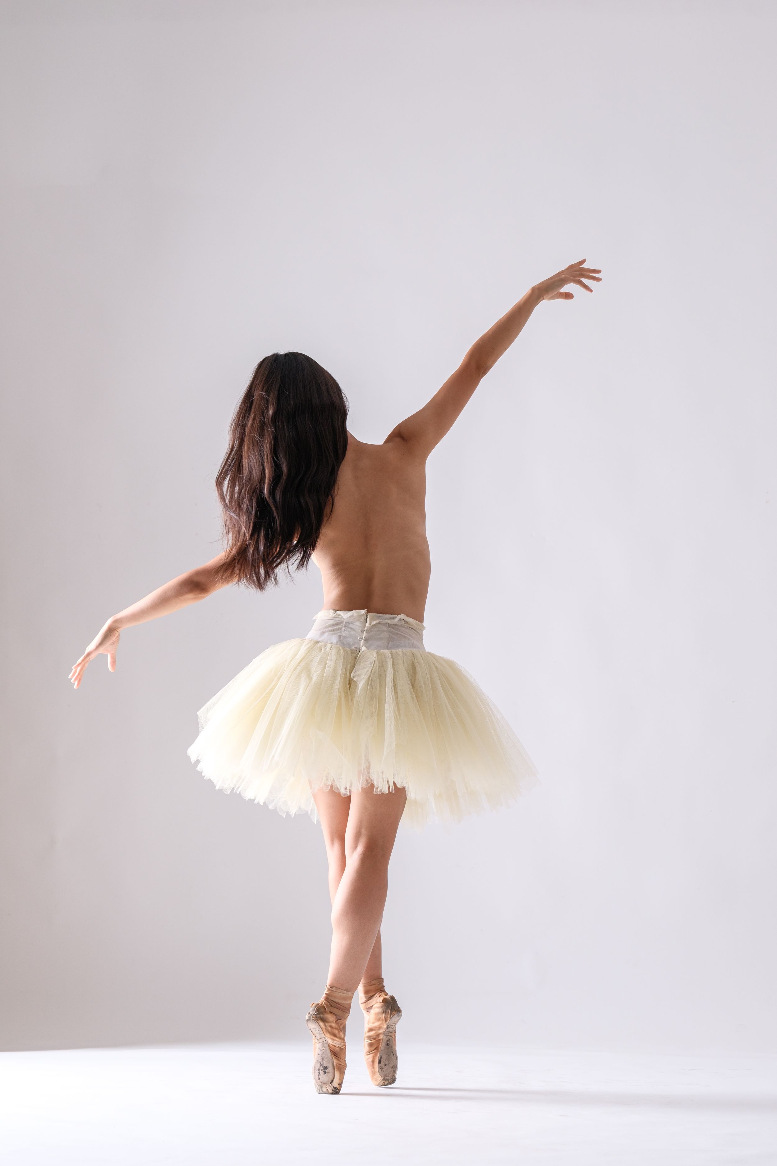 Dancer performs expressive movements in cream tutu and pointe shoes against white background.