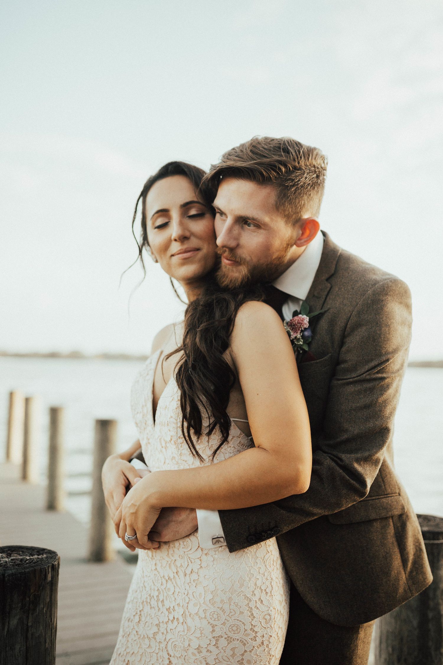 Bearded groom in a brown tweed suit hugs his bride in a lace wedding dress from behind on a waterfront wooden dock.