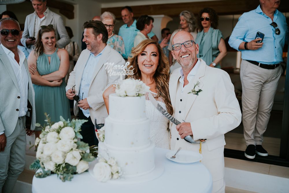 Wedding couple celebrates with guests while cutting their elegant white cake at outdoor reception.