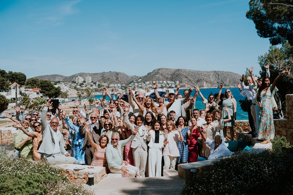 Large wedding party celebrates on hillside overlooking Mediterranean coastal views.