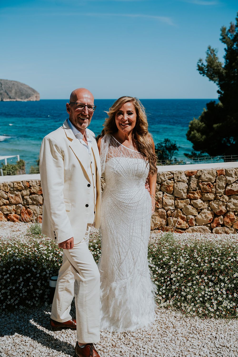 Newlyweds pose in white attire against stunning Mediterranean coastal backdrop with stone wall.