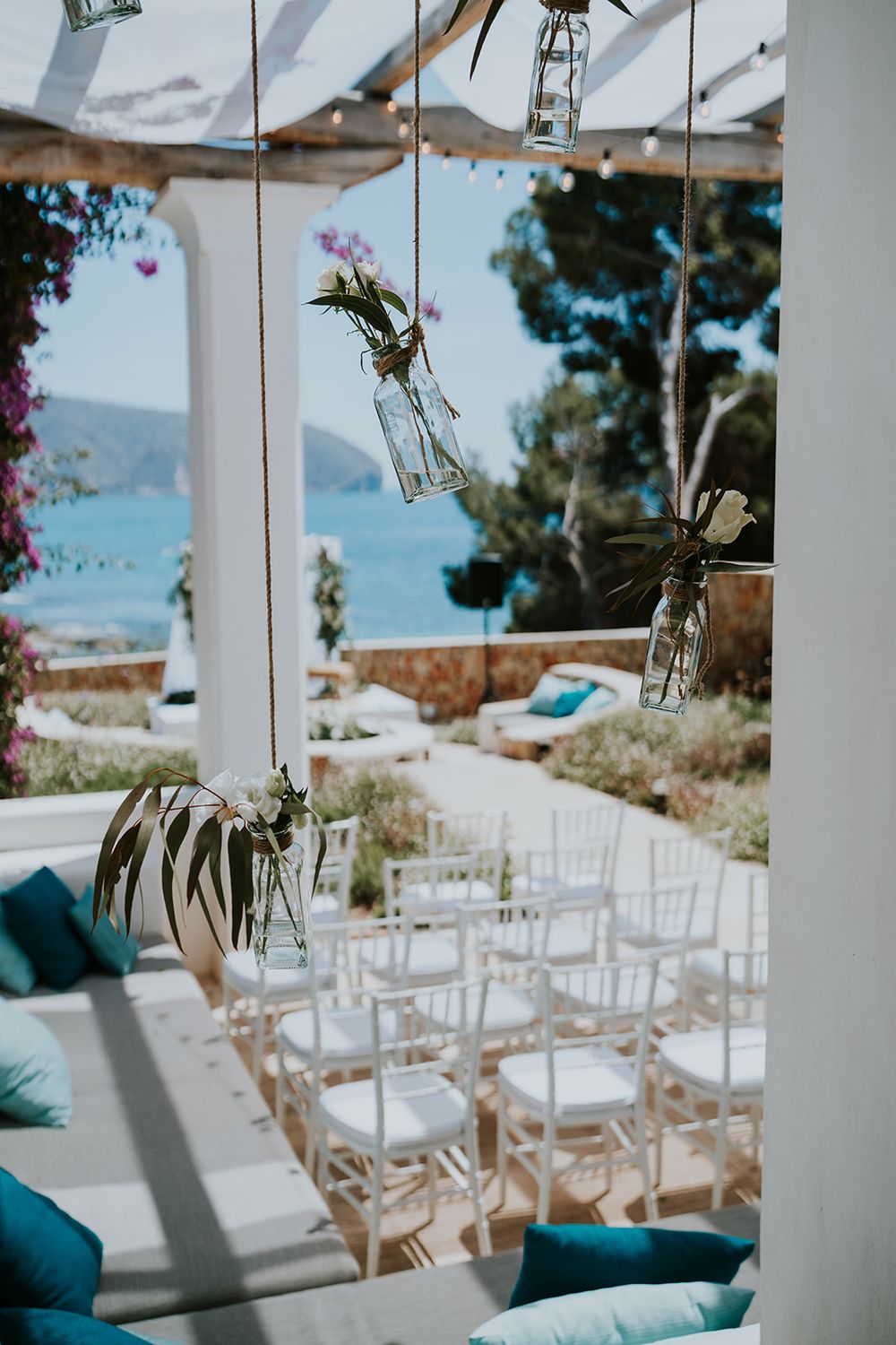 Elegant wedding ceremony setup with hanging glass vases and white chairs overlooking a beautiful ocean view.