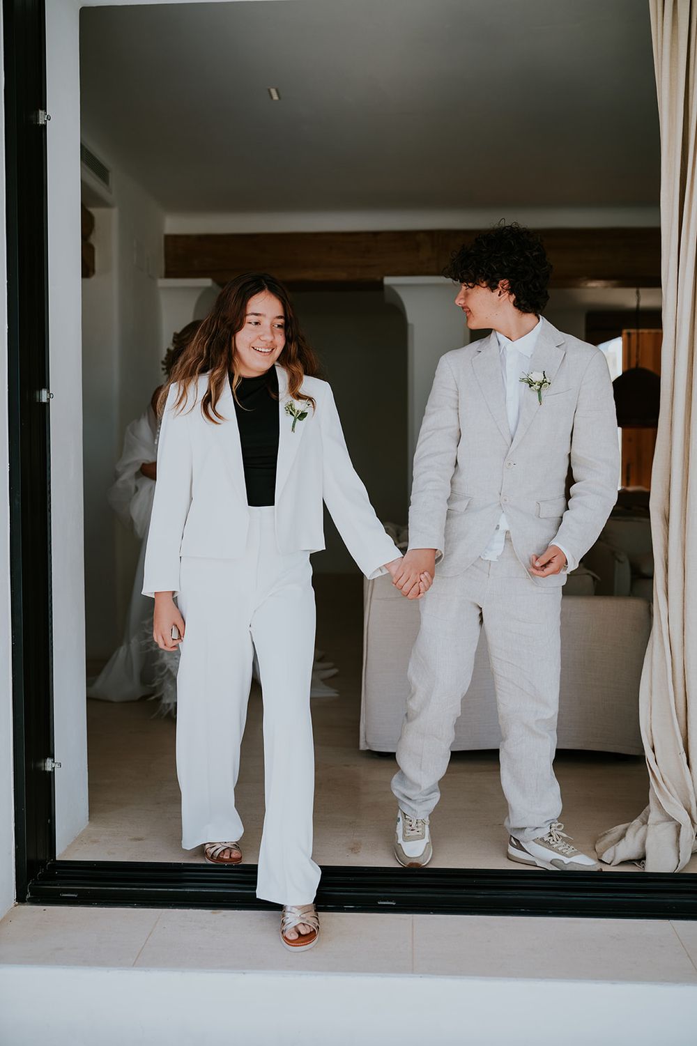 A couple in matching white suits hold hands while walking through an interior space.