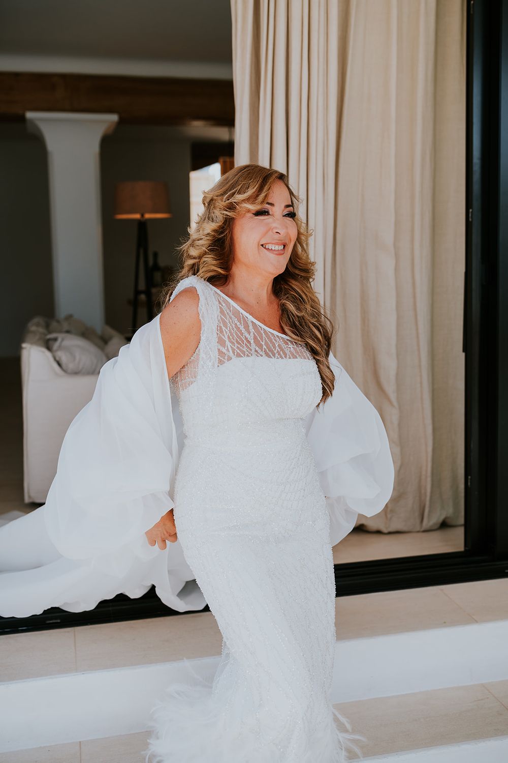A bride wearing a white lace dress with dramatic balloon sleeves smiles in an elegant interior.