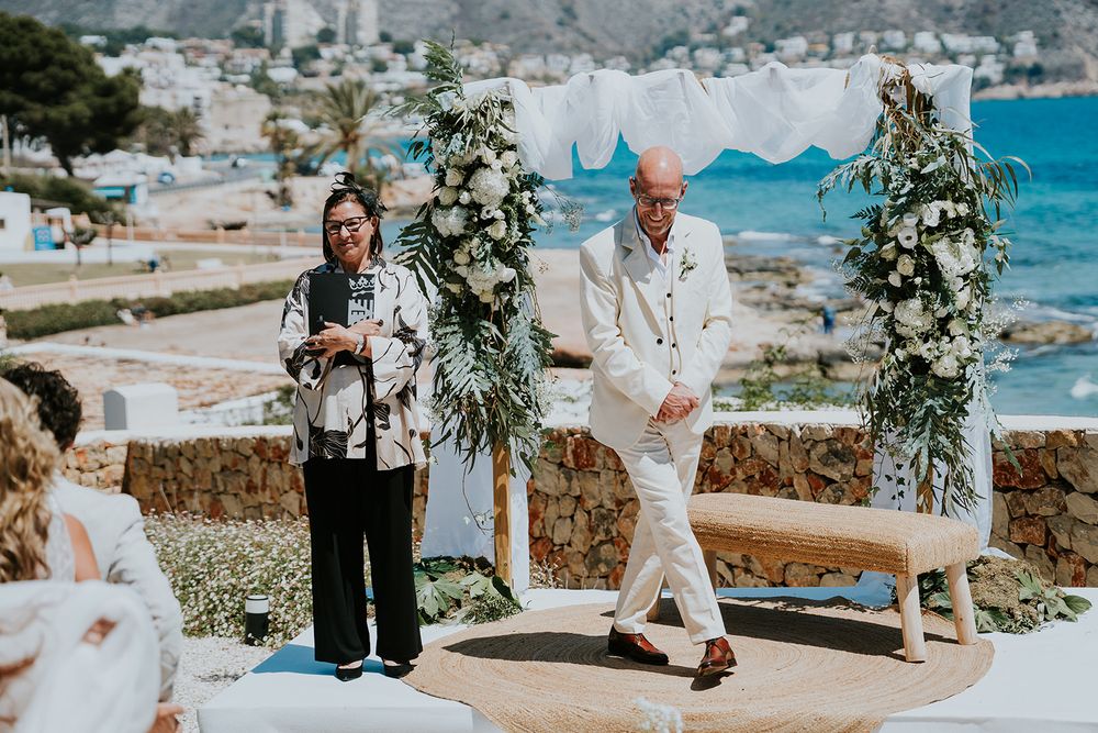 A seaside wedding ceremony setup with white floral arrangements and stone wall backdrop.