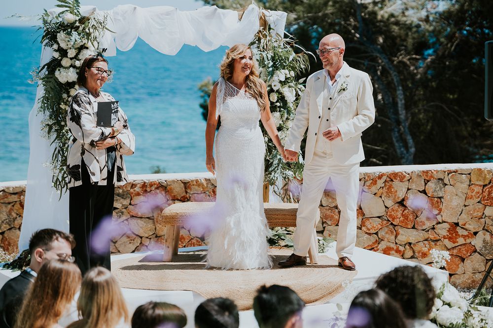 A wedding ceremony overlooking a blue ocean with floral arch and stone wall details.