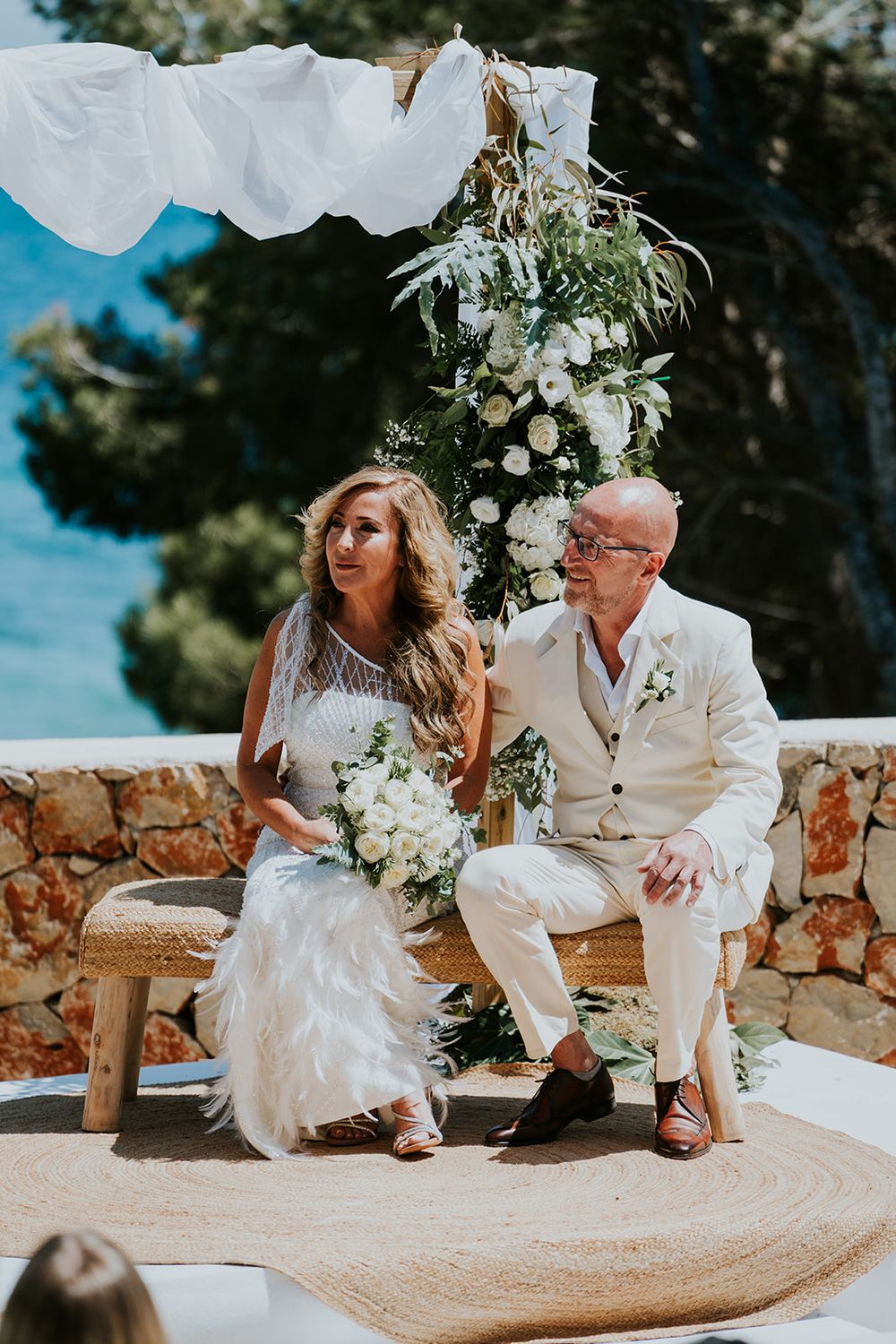 A couple sits together under a floral arch at their oceanfront wedding ceremony.