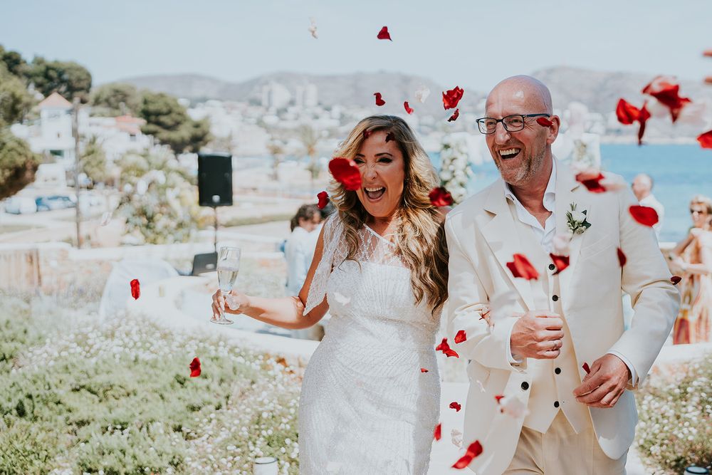Wedding celebrants share a candid moment of laughter as red petals shower down around them.
