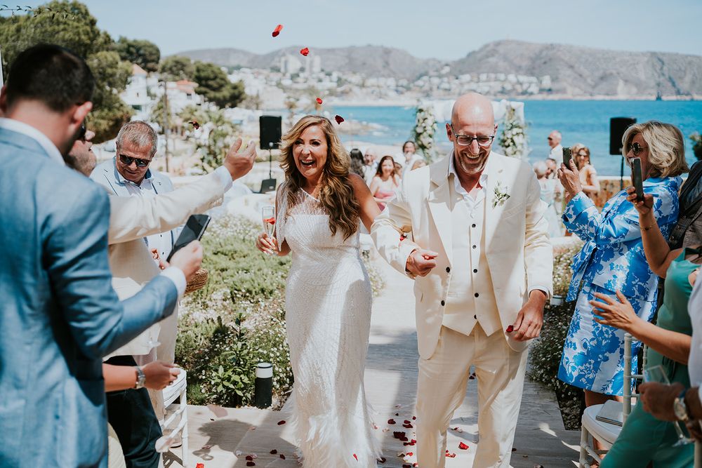 Wedding guests celebrate with the newlyweds as red petals fill the air at an outdoor coastal ceremony.