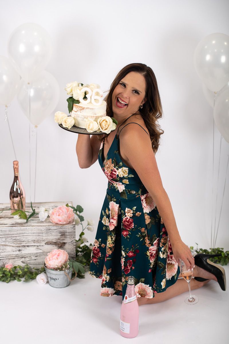 Celebration scene with floral dress, cake, champagne and white balloons in studio setting.