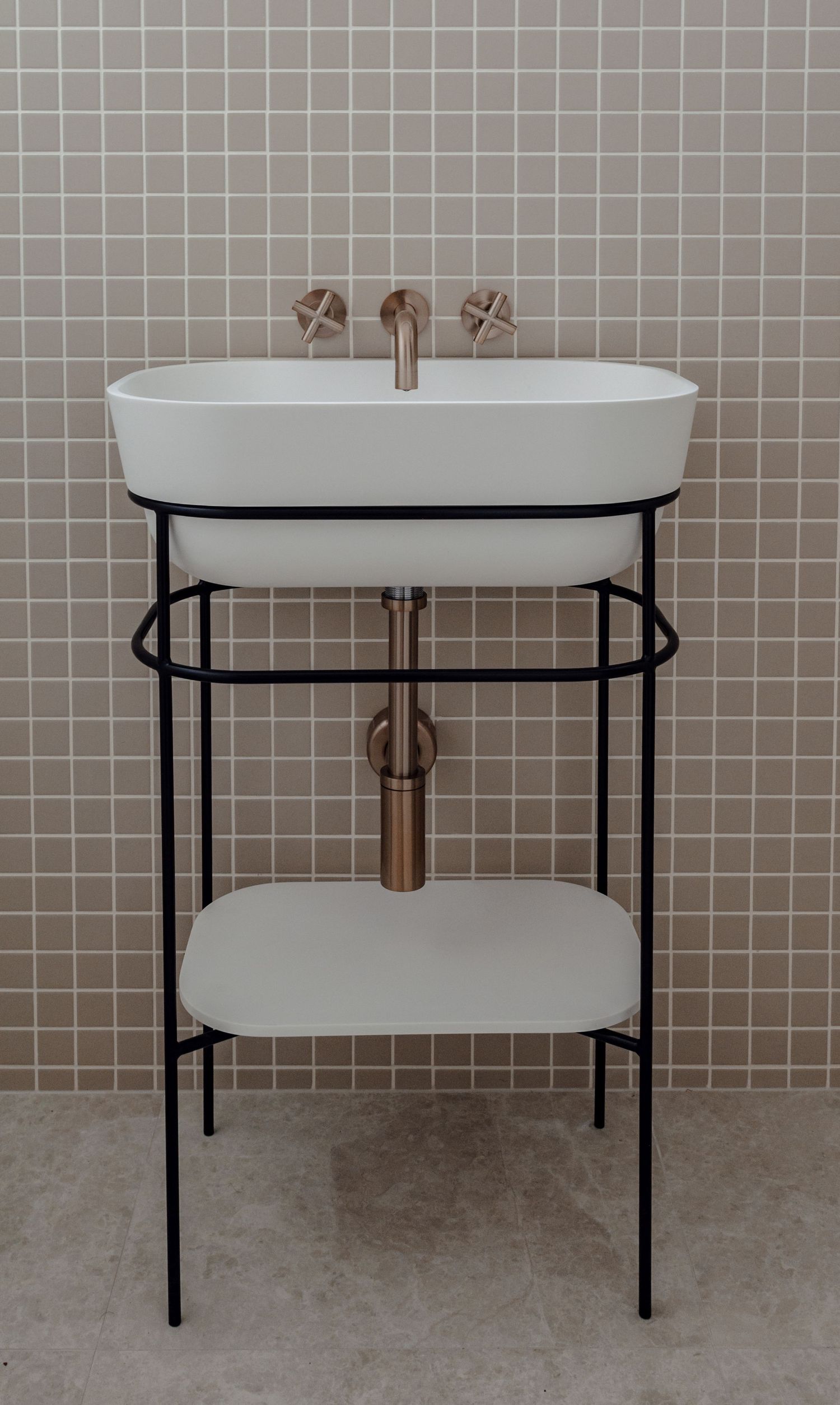 Modern bathroom vanity with white basin, black metal frame and brass fixtures against tiled wall.